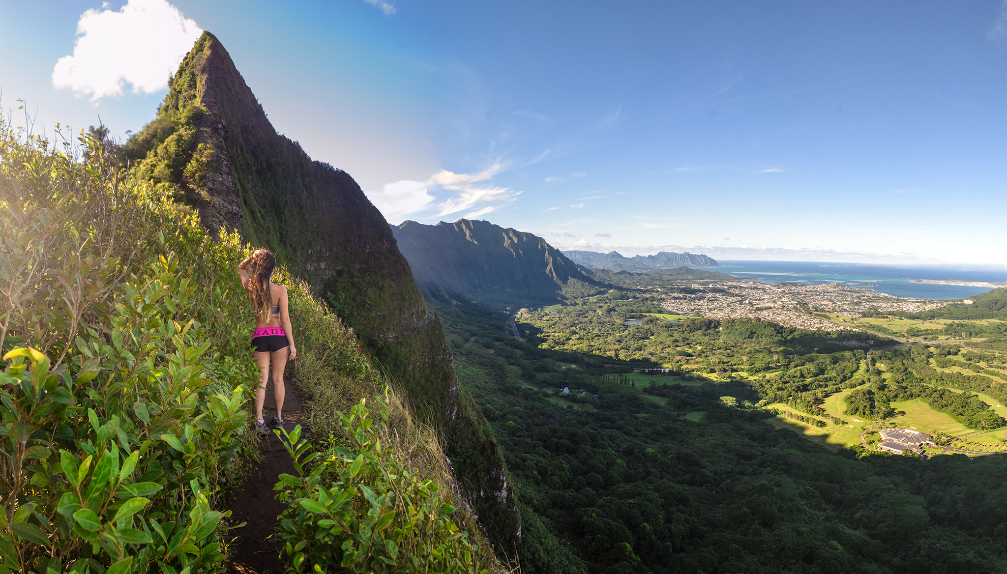 A hiker on the Nuuanu Saddle ridge trail on Oahu, overlooking lush valleys, jagged mountains, and the Pacific Ocean in the distance.
