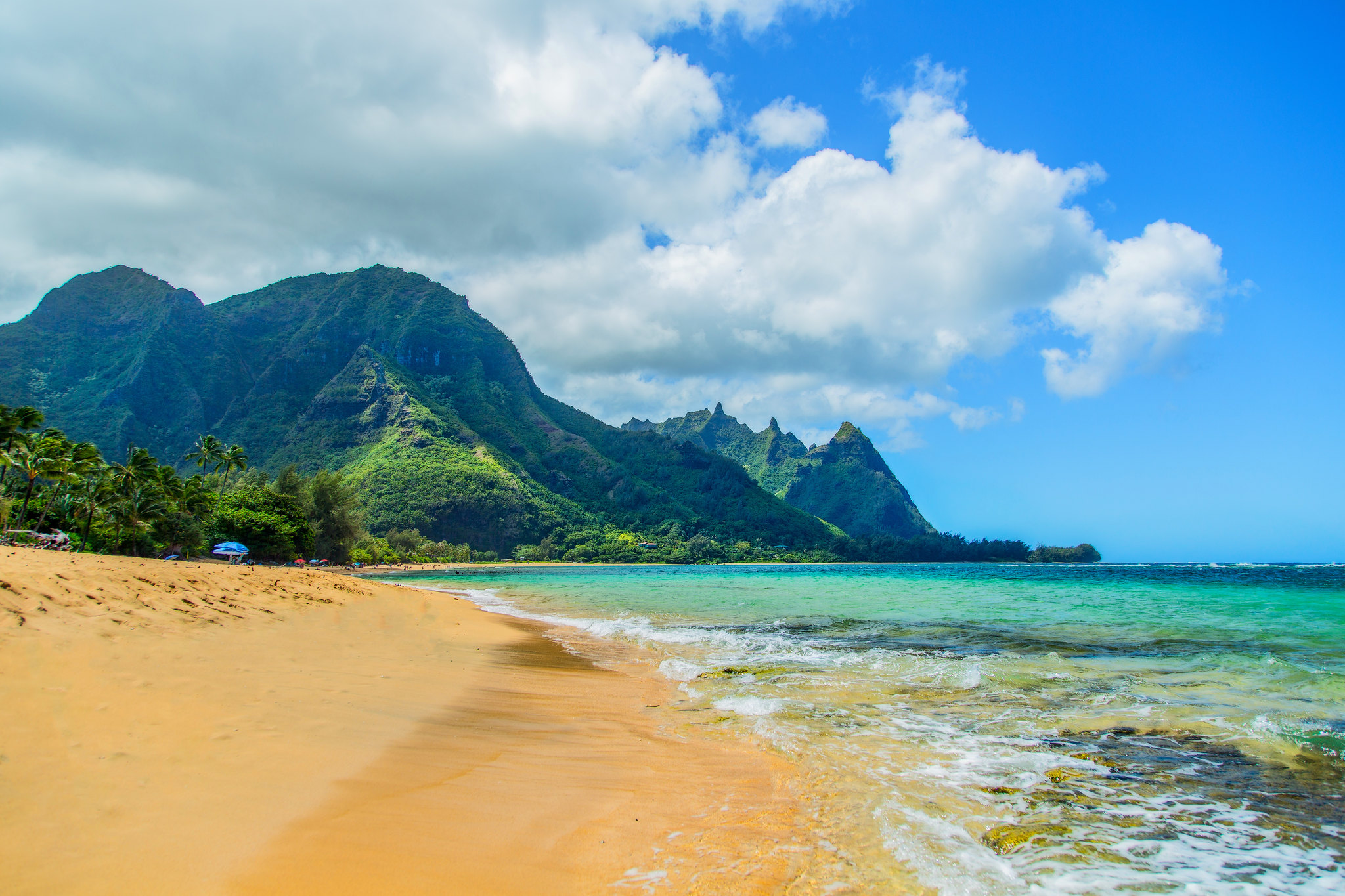 Golden sand, turquoise ocean, and lush green mountains at Ha'ena Beach on Kauai’s North Shore under a partly cloudy sky.