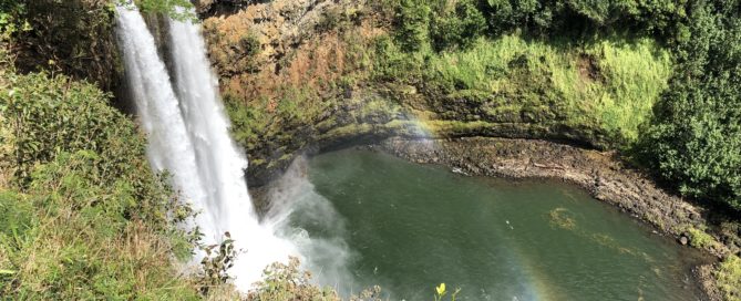 Wailua Falls on Kauai, Hawaii, with water plunging into a green pool below. A faint rainbow appears in the mist, surrounded by lush tropical foliage.