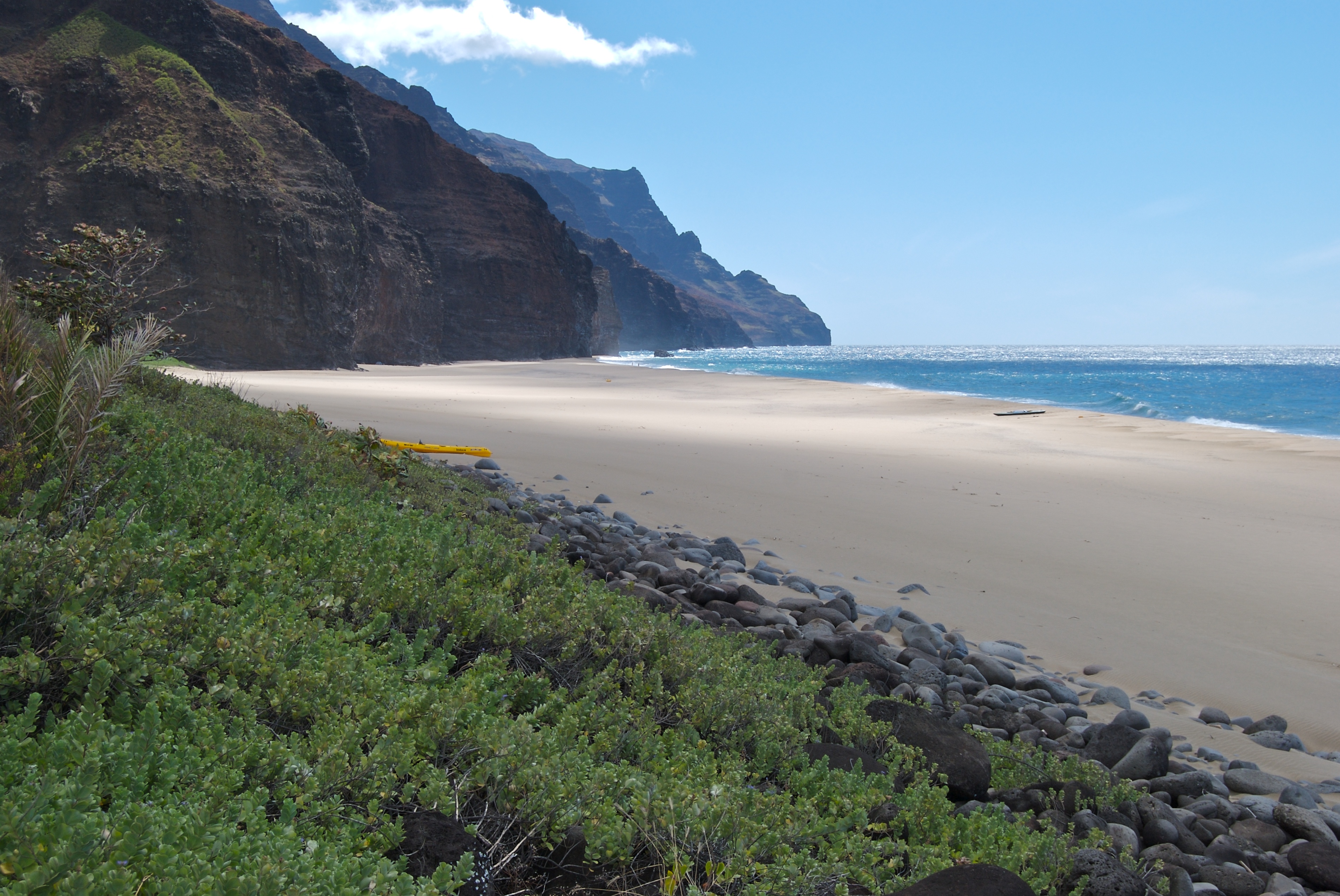 Yellow kayak resting on the sandy shore of the dramatic Napali Coast, Kauai, with turquoise water and lush green cliffs in the background.